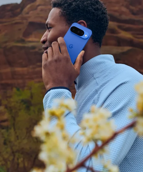 A man using a blue Google Pixel 10 outdoors, with its rear camera bar visible against a rocky landscape.